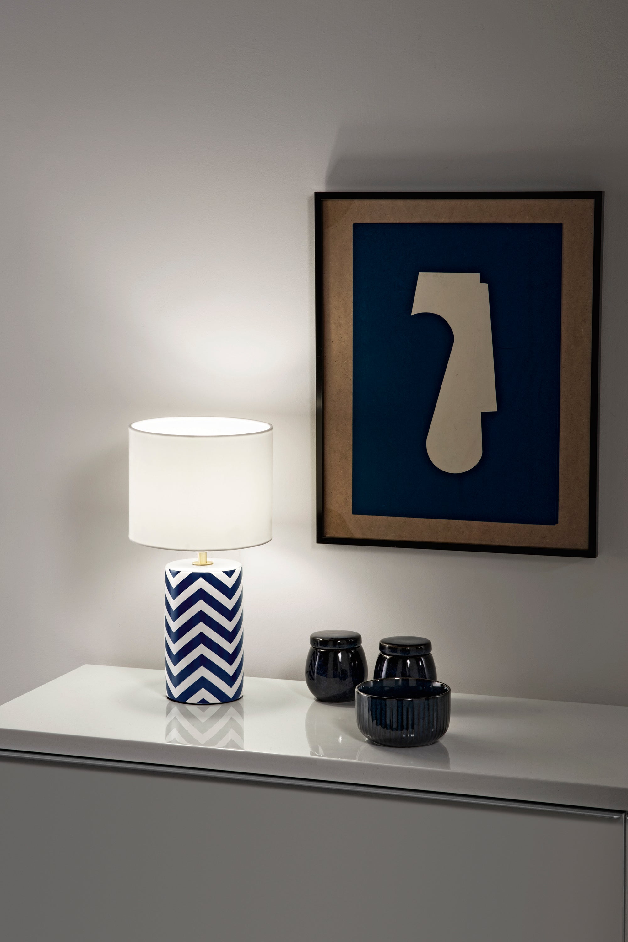 Lampada da tavolo Zeno accesa a righe blu e bianche su consolle moderna che illumina un quadro